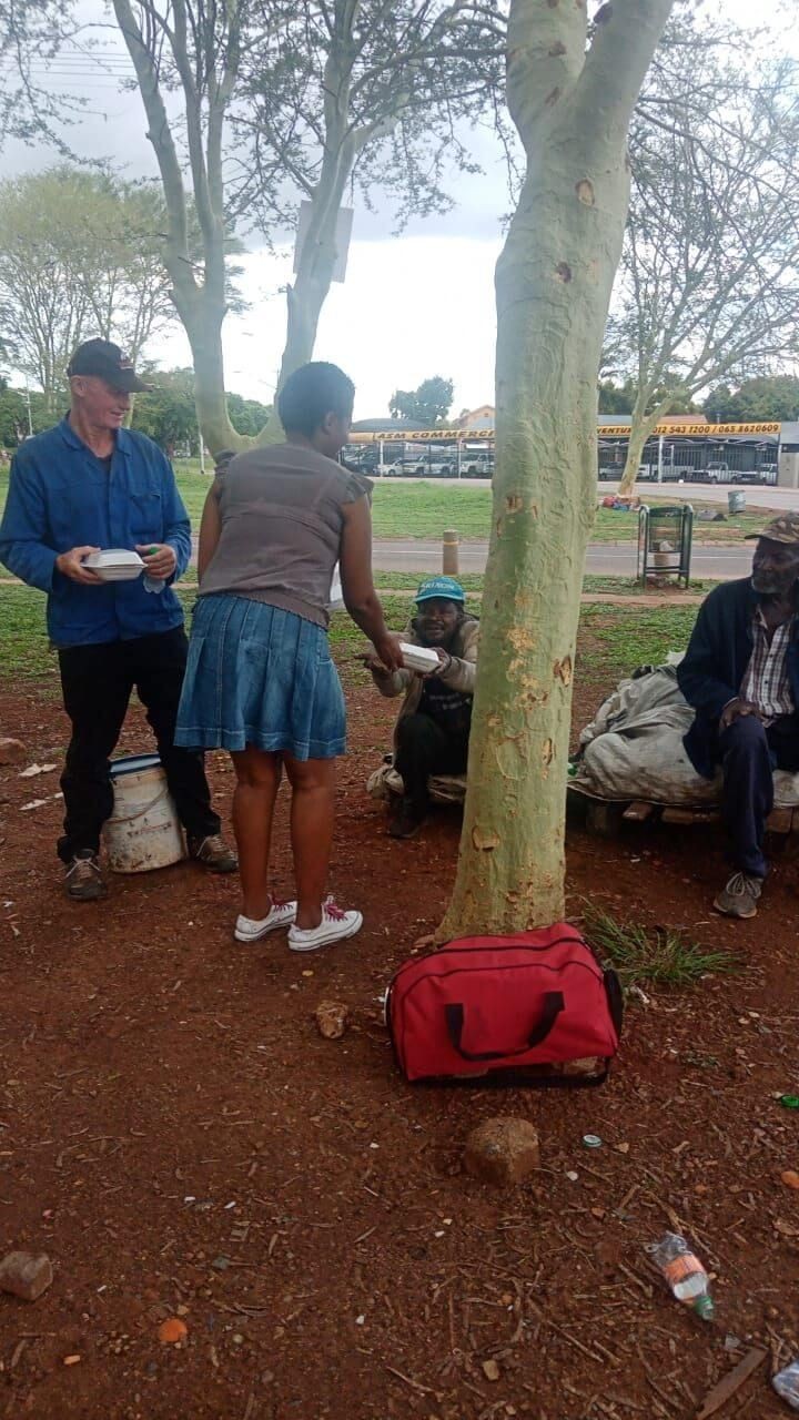 Person handing food containers to seated individuals in a park, with trees and a red bag nearby.