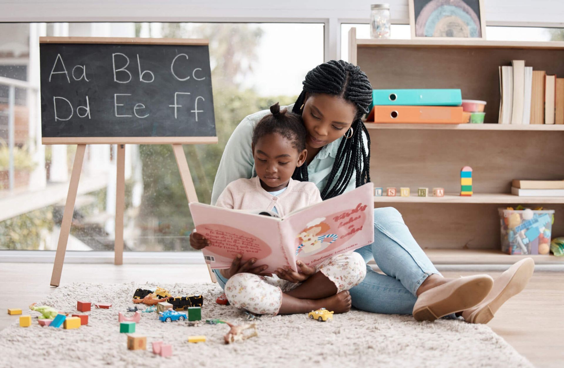 Woman and child reading a book together in a playroom with toys and alphabet chalkboard.